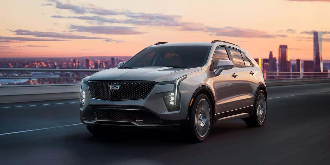 Silver SUV driving on a bridge at sunset with a city skyline in the background.