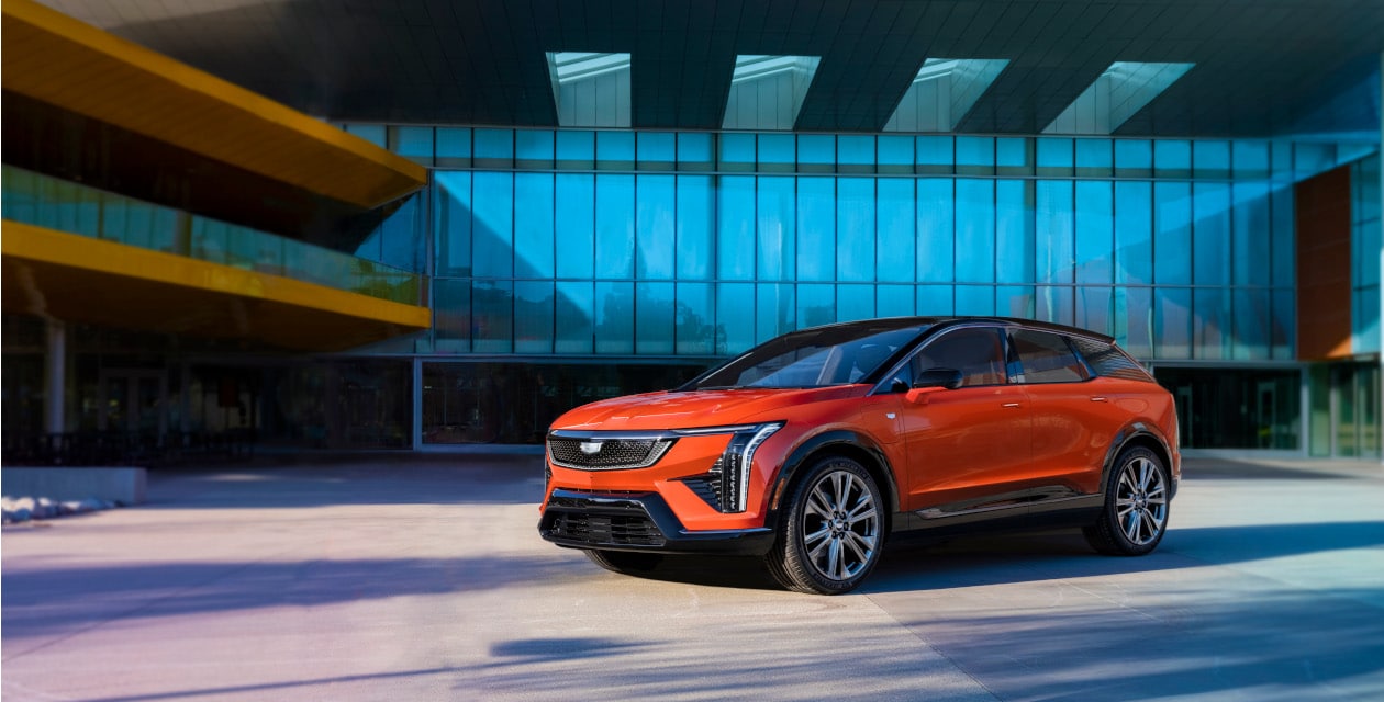 Orange SUV parked in front of a modern glass building, casting a clear shadow.