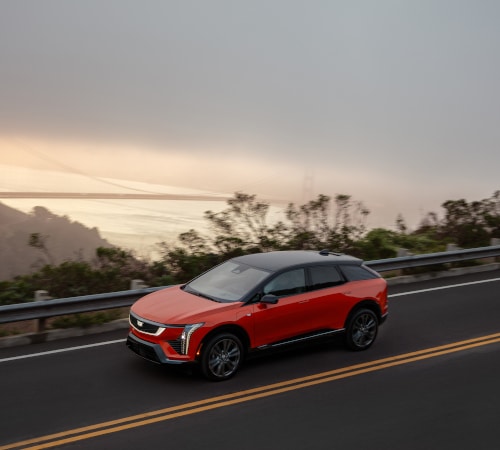 Red SUV driving on a coastal road with foggy hills and sunrise in the background.