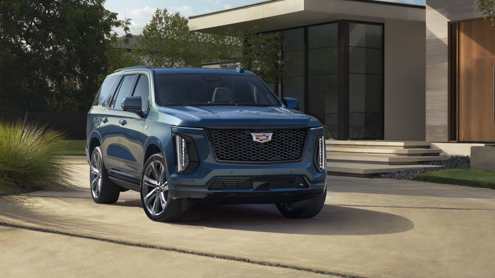 Blue Cadillac SUV parked in front of a modern house, surrounded by lush greenery.