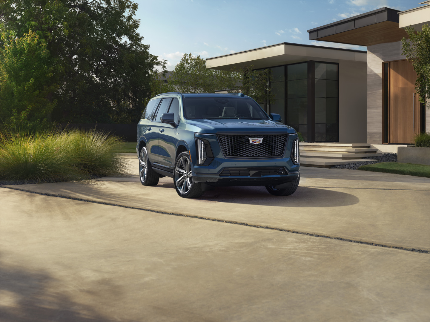 Blue Cadillac SUV parked in a modern driveway with trees and a contemporary house in the background.